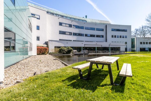 Zonnig buitenterrein met picknicktafels, vijver en het moderne gebouw van Sparrenheuvel op de achtergrond.