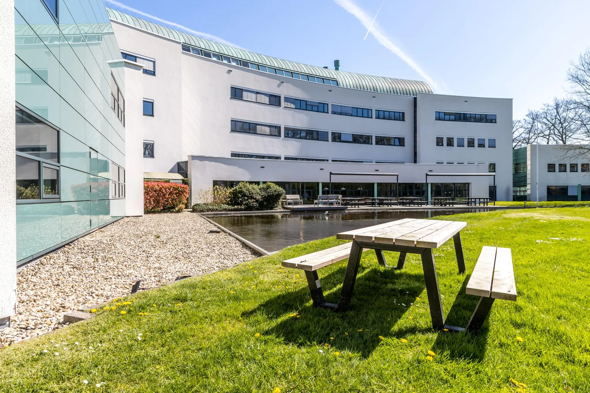 Zonnig buitenterrein met picknicktafels, vijver en het moderne gebouw van Sparrenheuvel op de achtergrond.