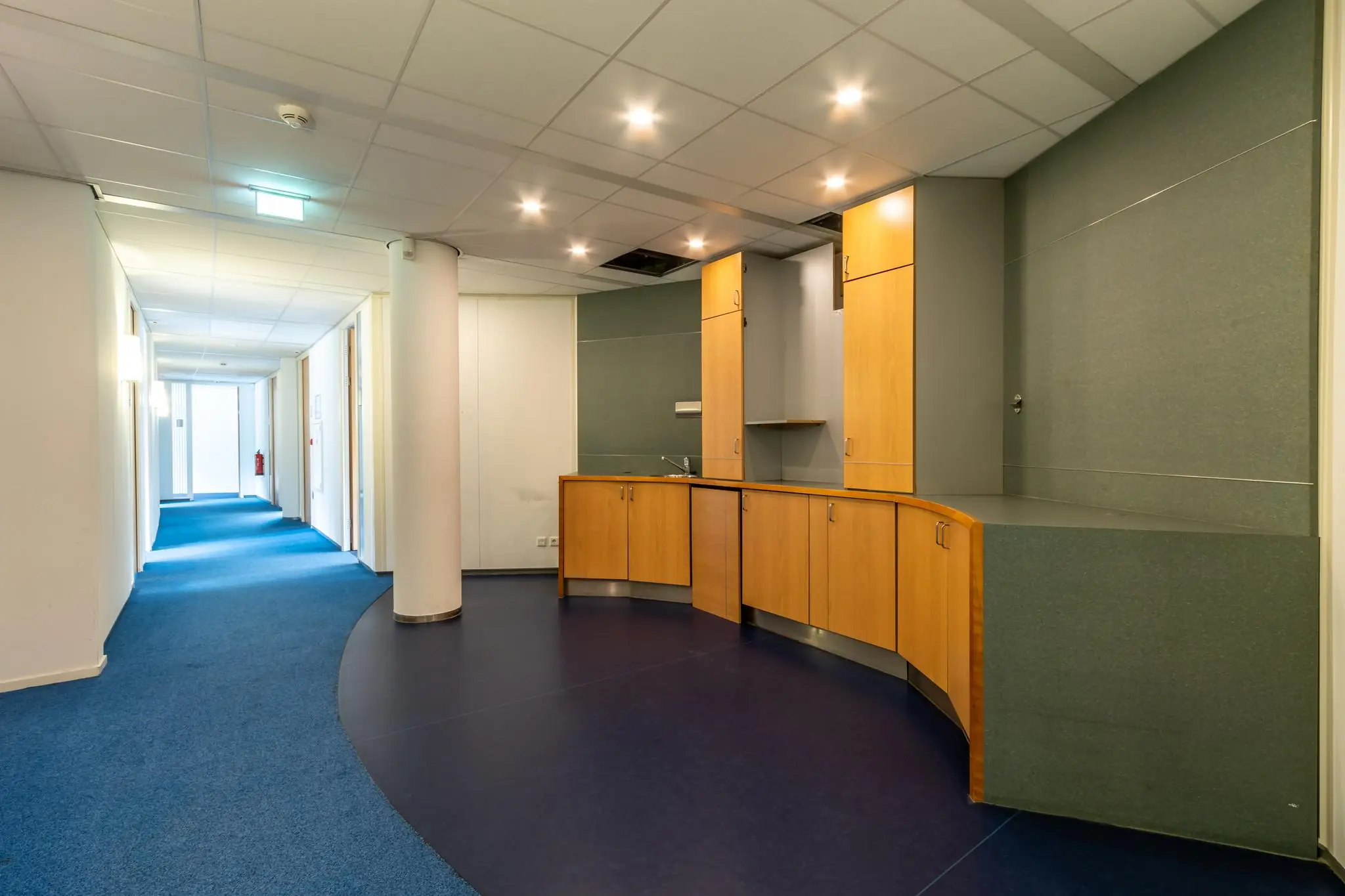 Hal met blauwe vloerbedekking en ingebouwde houten pantry in kantoorgebouw Sparrenheuvel.