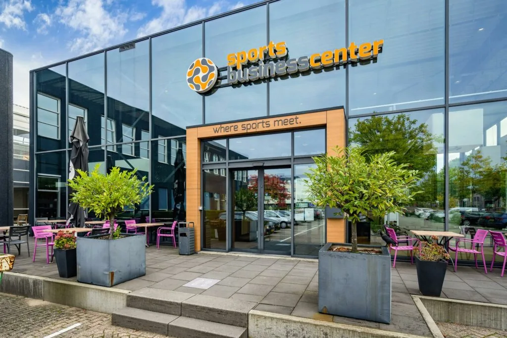 Moderne glazen gevel van het Sports Business Center aan de Plesmanstraat met terras en kleurrijk meubilair.