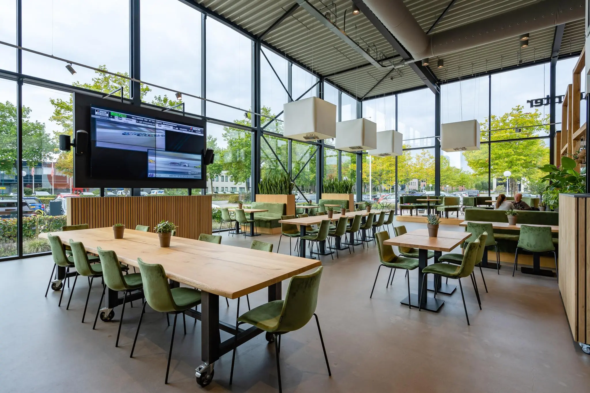 Moderne en licht ingerichte kantine aan de Plesmanstraat met groene stoelen, houten tafels en grote ramen met uitzicht op bomen.