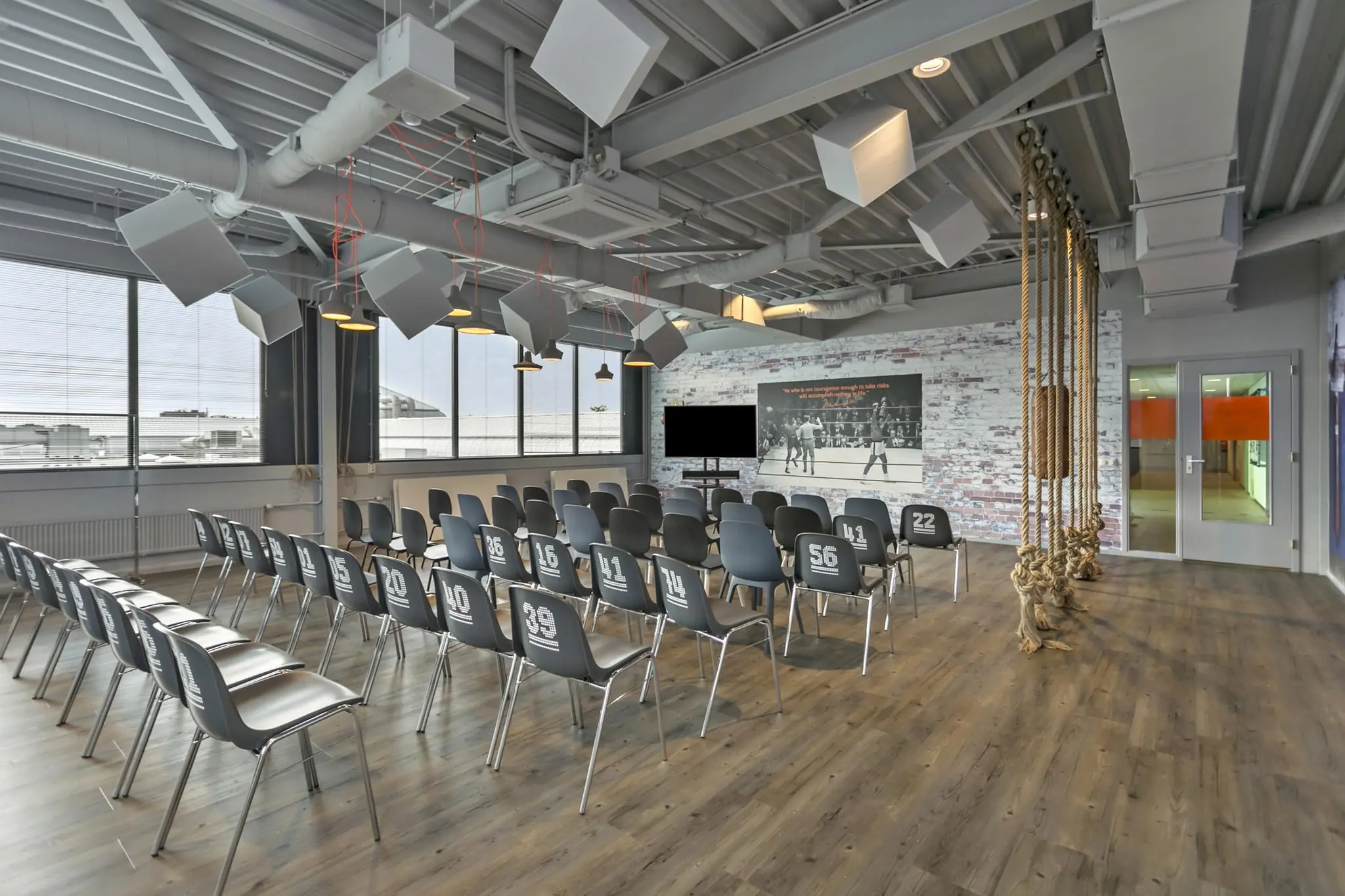 Moderne presentatieruimte aan de Plesmanstraat met stoelen, tv-scherm, bokswanddecoratie en industriële inrichting.