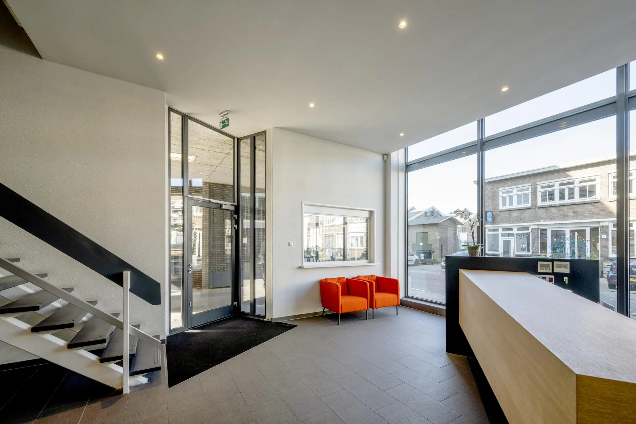 Moderne entreehal met grote ramen, een houten balie, twee feloranje stoelen en een trap naar boven aan de Helicopterstraat.