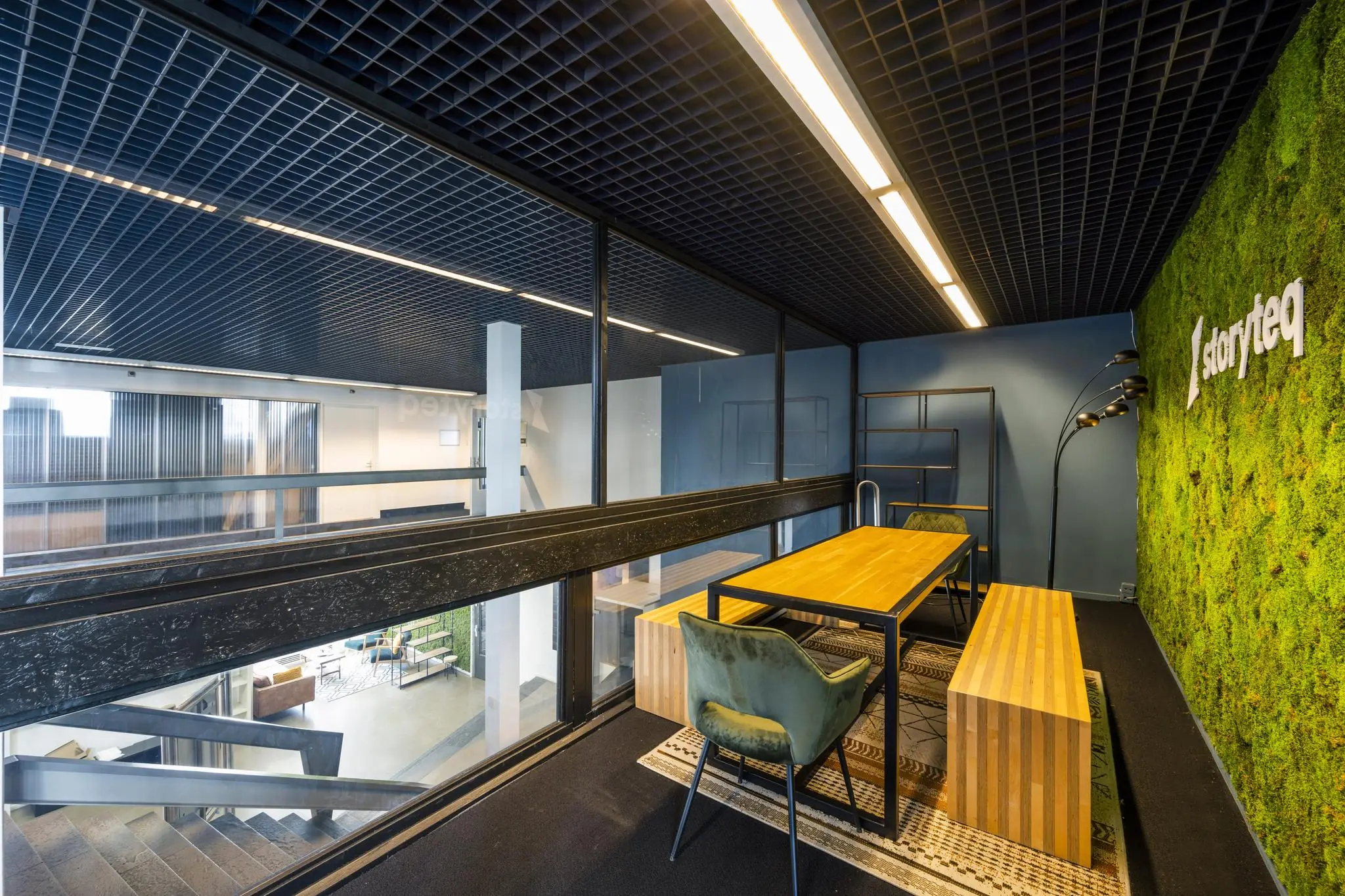 Modern kantoorinterieur met houten tafel, groene moswand met "Storyteq"-logo en uitzicht op een open trappenhal.