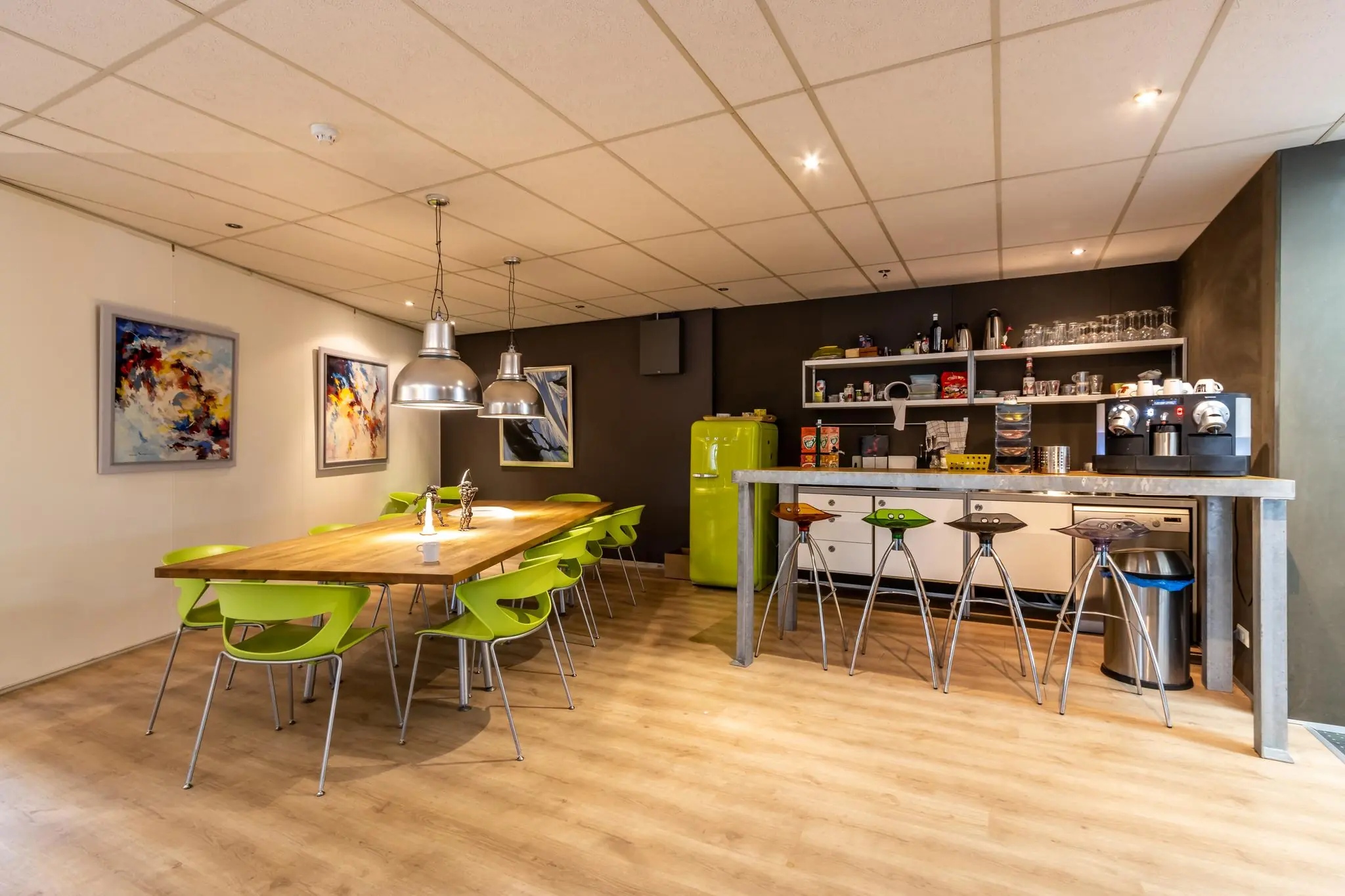 Moderne kantine met een lange houten tafel, groene stoelen, kleurrijke kunst aan de muur en een open keuken met koffieapparaat en felgroene retro koelkast.