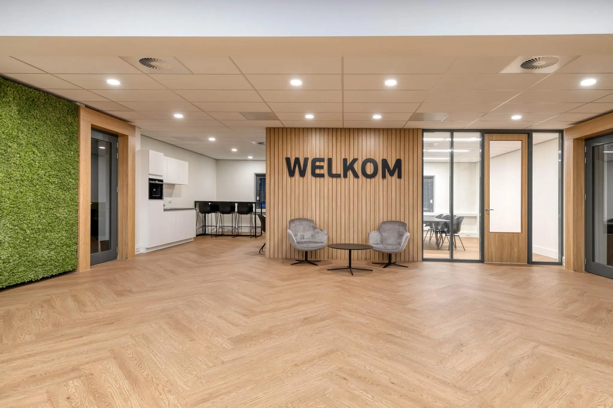 Moderne ontvangsthal met houten vloer, groene moswand en de tekst "WELKOM" op een houten wand.