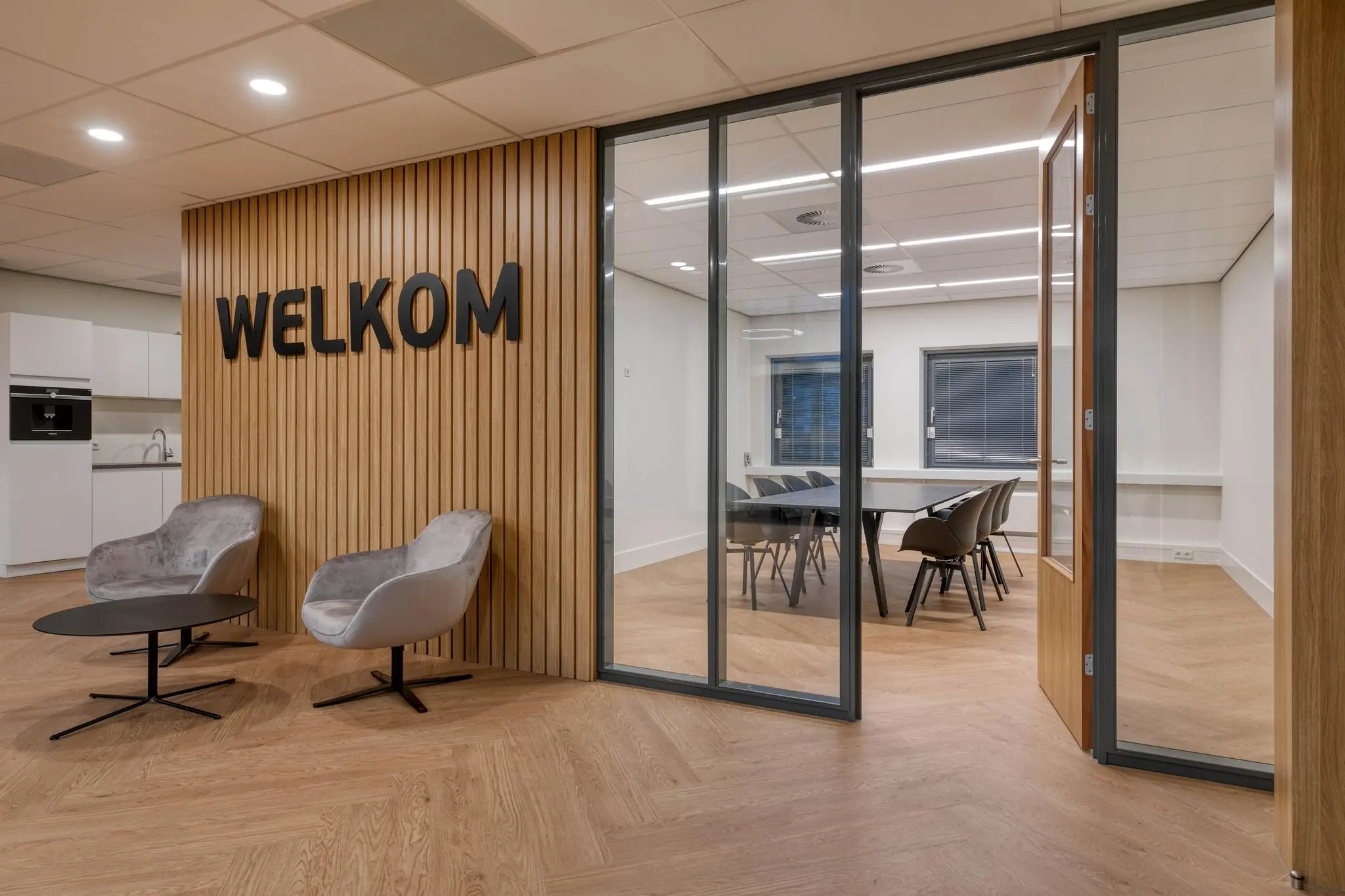 Moderne kantoorentree met houten wand met het woord “WELKOM”, twee fauteuils, en zicht op een vergaderruimte met glazen wanden.