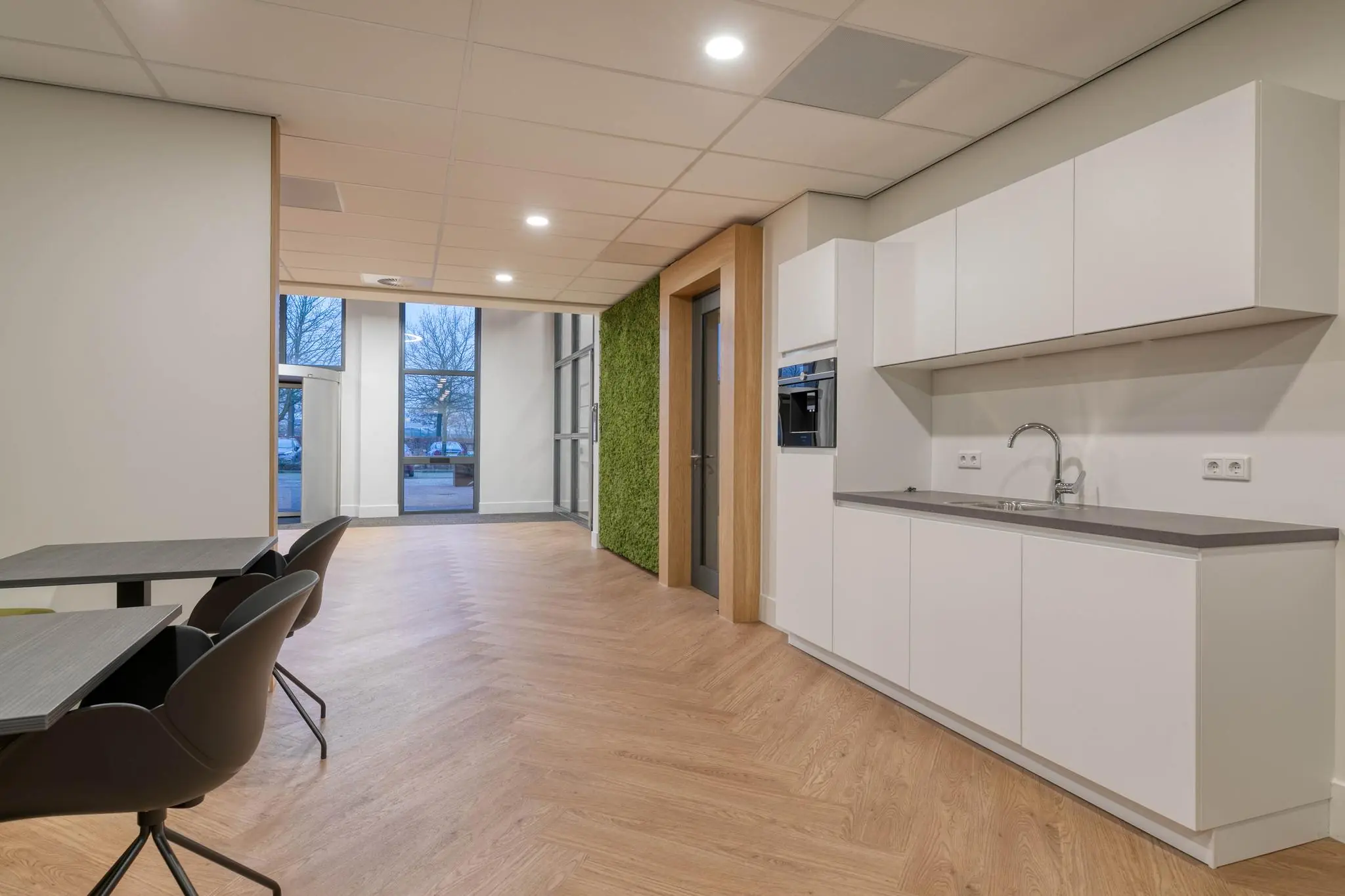 Moderne kantoorkeuken met witte kasten, ingebouwde koffiemachine en zitgedeelte met uitzicht op buiten.