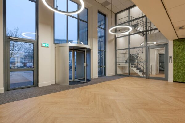 Moderne, lichte entreehal van een kantoorgebouw met houten visgraatvloer, glazen wanden en ronde plafondverlichting.