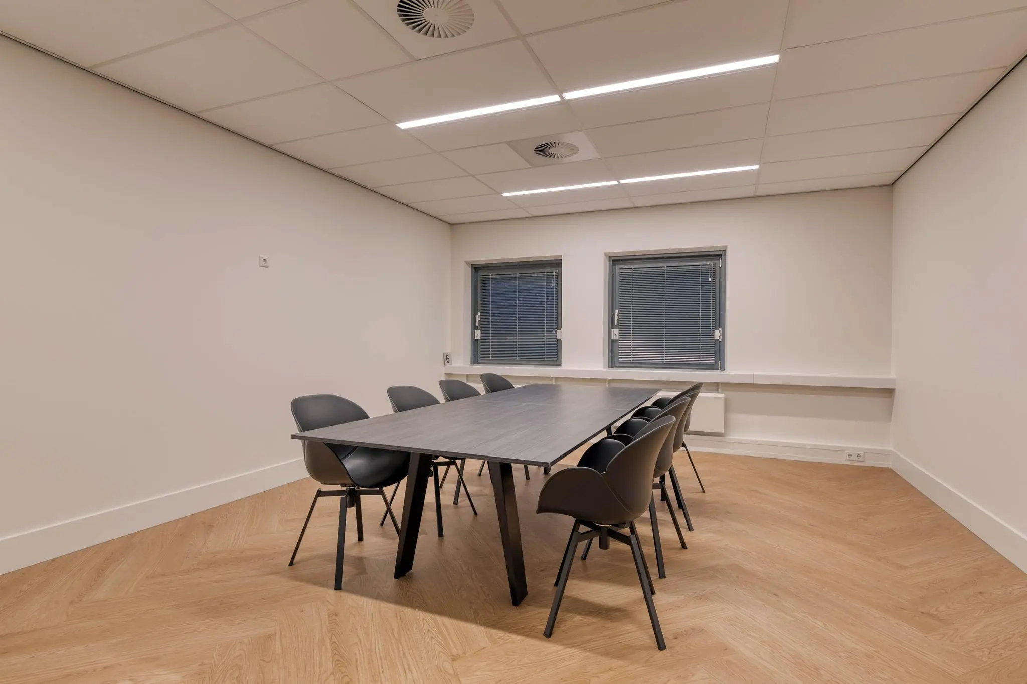 Vergaderruimte met een lange tafel, acht zwarte stoelen en twee ramen met jaloezieën.