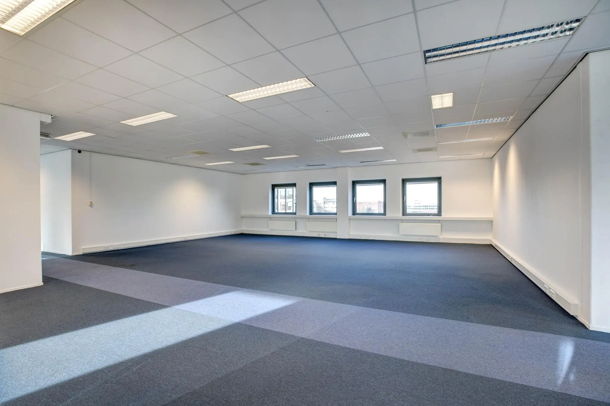 Licht en ruim leeg kantoor met blauw tapijt, systeemplafond en drie ramen met uitzicht op de stad.