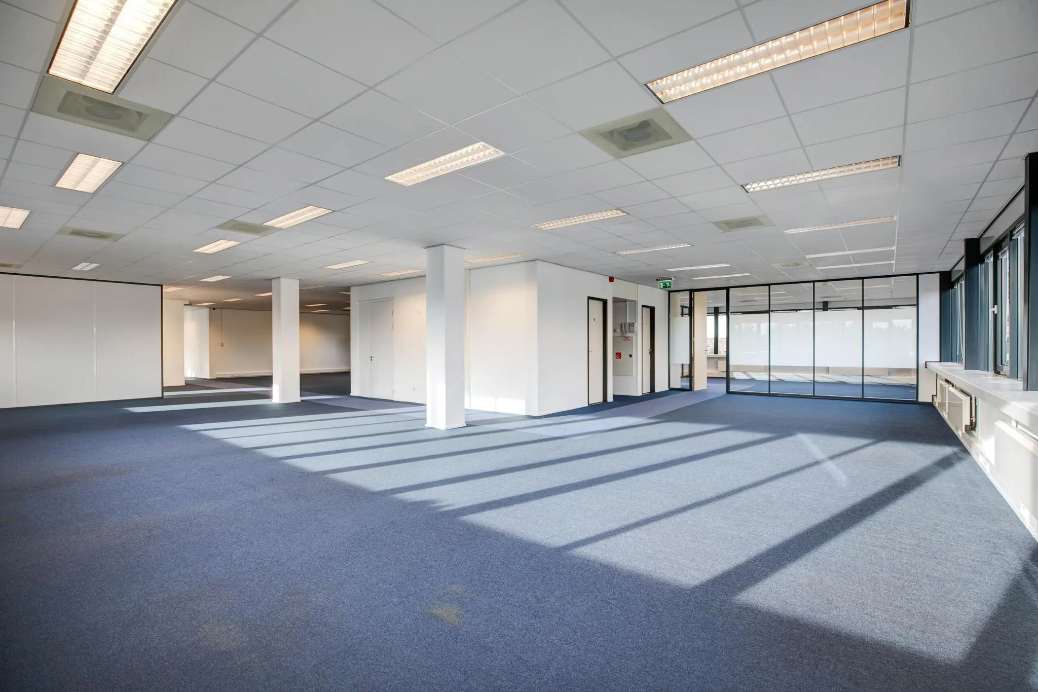 Lichte, lege kantoorruimte met blauw tapijt, systeemplafond en grote ramen.
