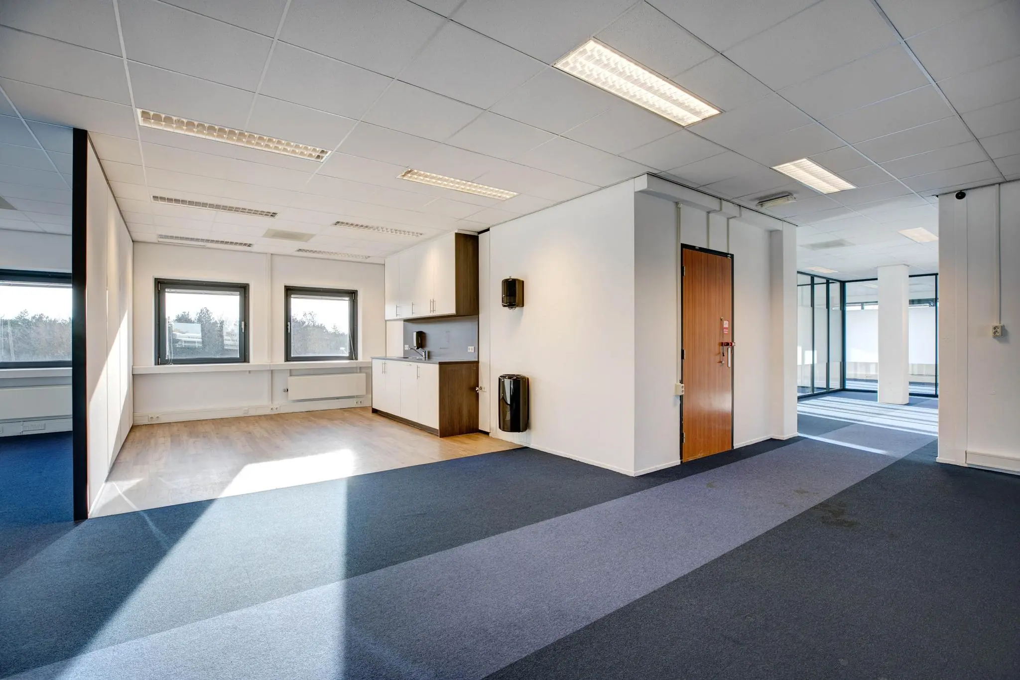 Moderne, lichte kantoorruimte met keukenblok, blauwe vloerbedekking en grote ramen.