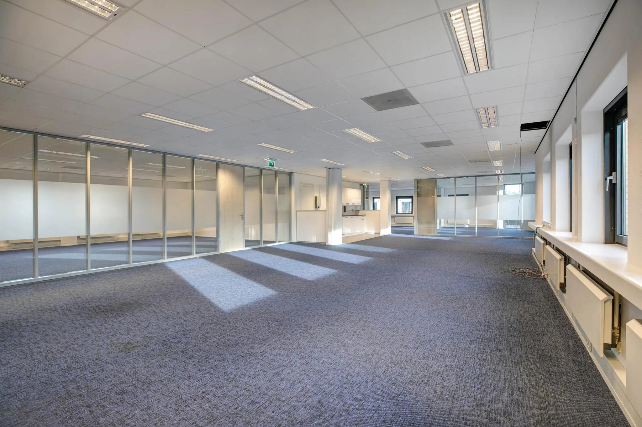 Ruime, leegstaande kantoorruimte met glazen scheidingswanden en blauwe vloerbedekking.