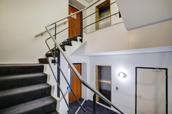 Interieur van een trappenhuis met een metalen trapleuning, tapijttrap, liftdeuren en houten deuren.