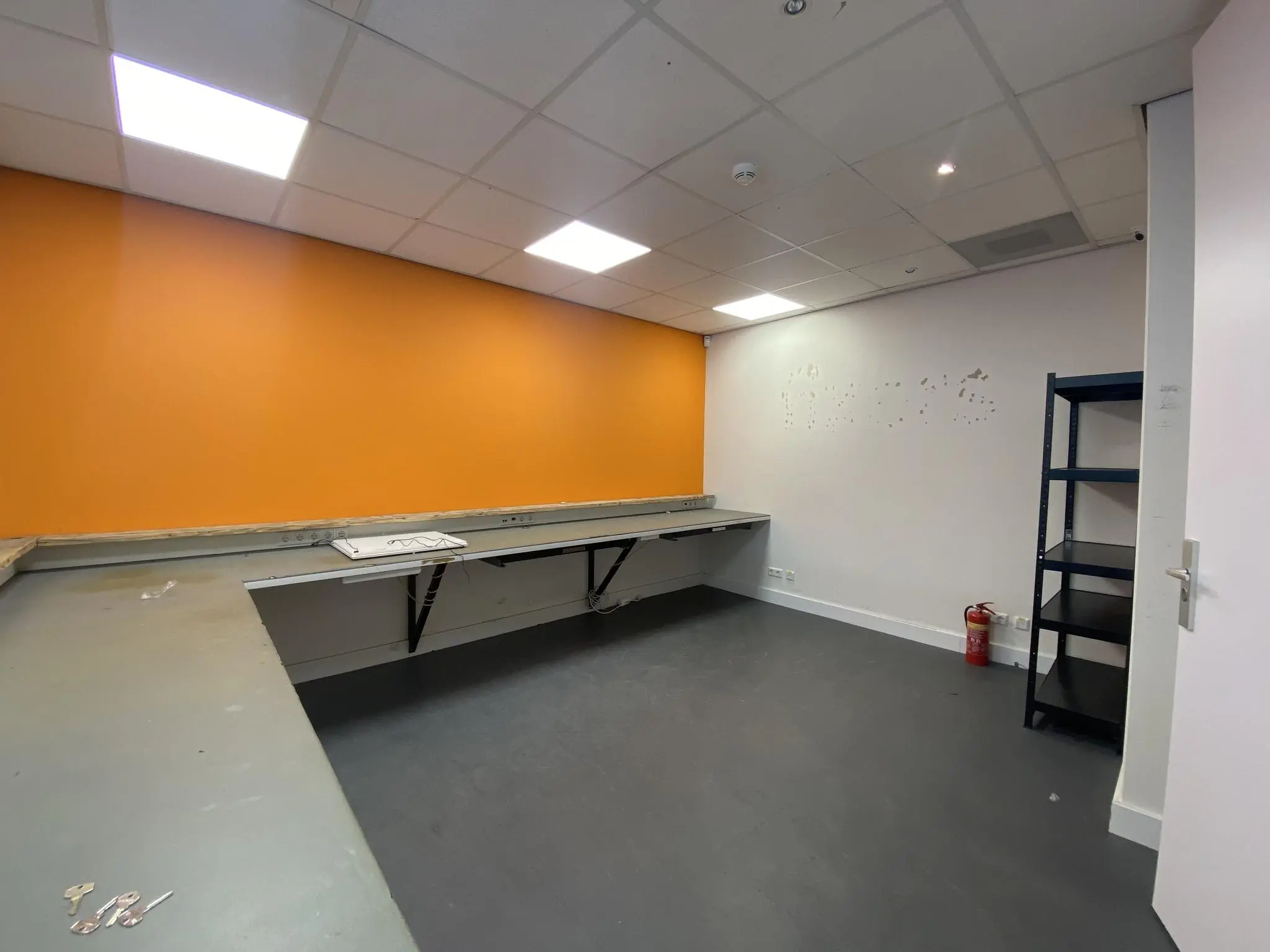 Lege kantoorruimte aan het Herculesplein met oranje accentmuur, lange werktafel, metalen stellingkast en een brandblusser.