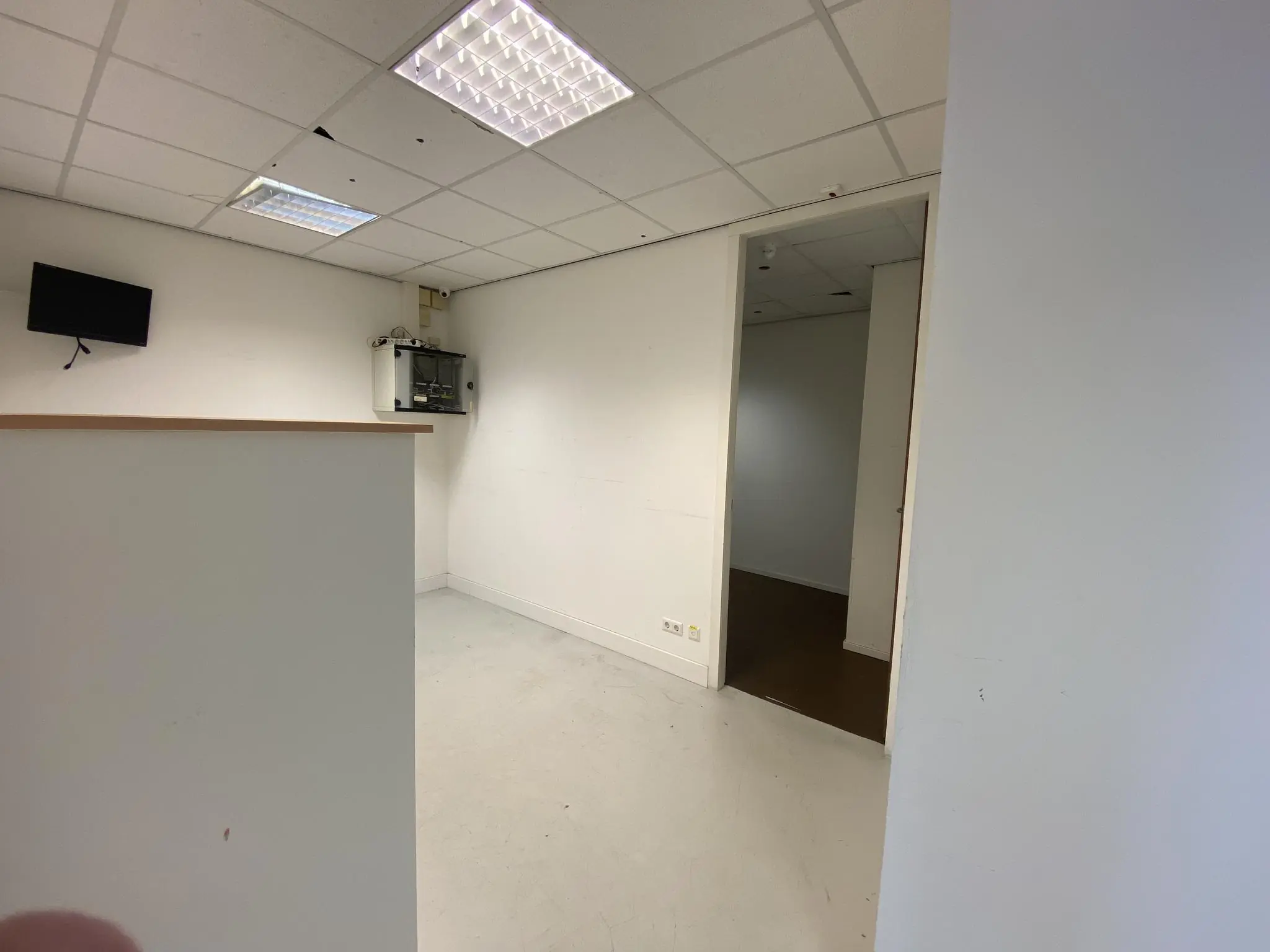Een lege, witte kantoorruimte op Herculesplein met systeemplafond, wandmonitor en openstaande deur naar een aangrenzende kamer.