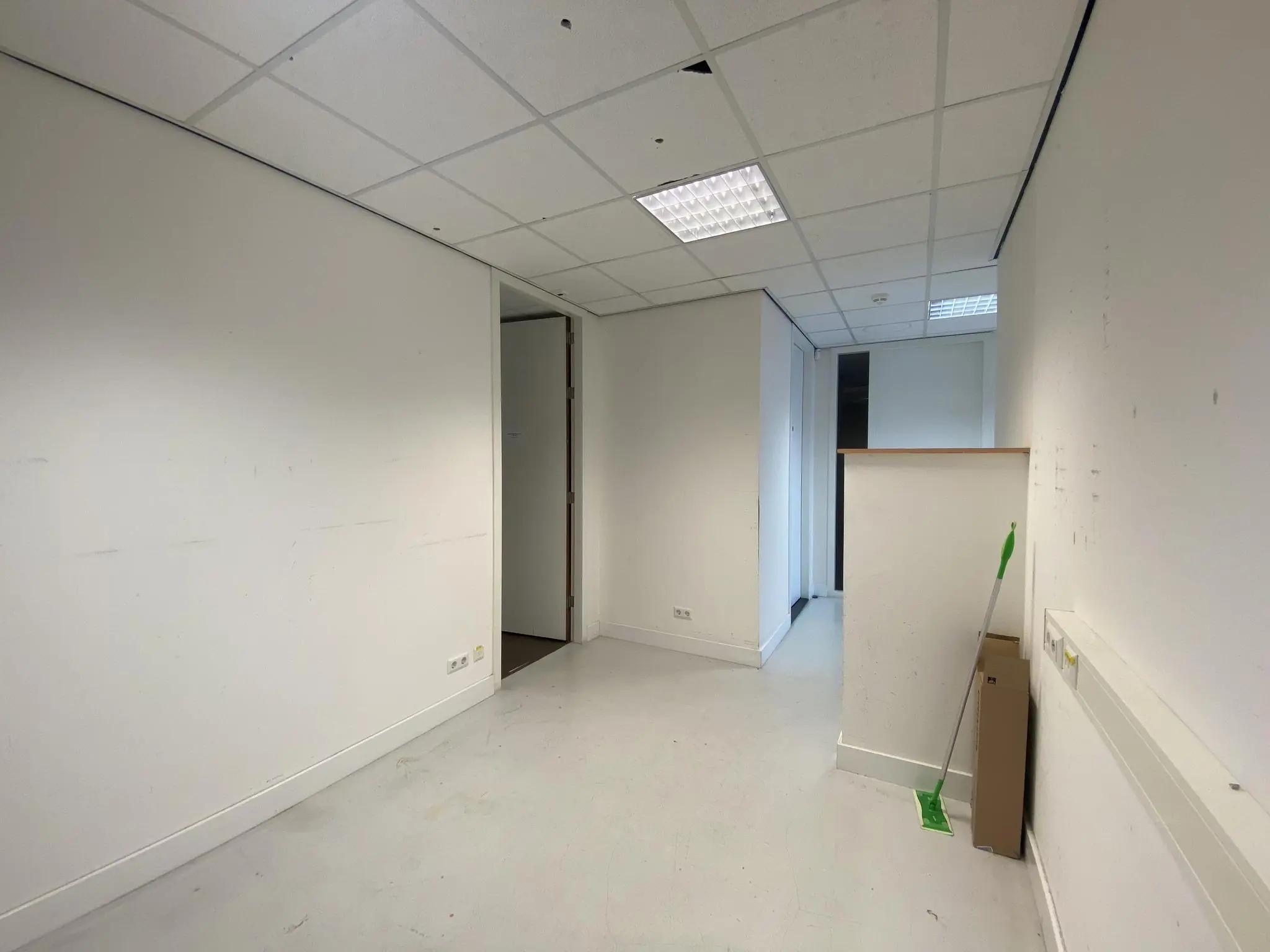 Een lege, witte kantoorruimte op het Herculesplein met systeemplafond en schoonmaakmateriaal in de hoek.