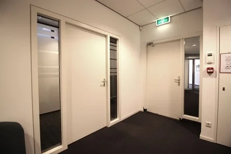 Hal van een kantoorgebouw aan het Herculesplein met witte deuren, glazen panelen en een nooduitgangsbord.
