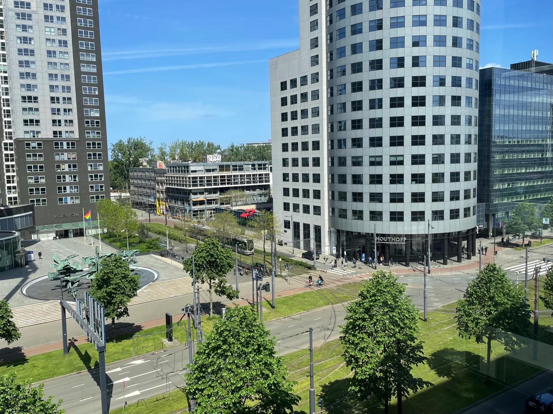 Zicht op het Weena in Rotterdam met moderne kantoorgebouwen, trambanen en een plein met kunstinstallatie.