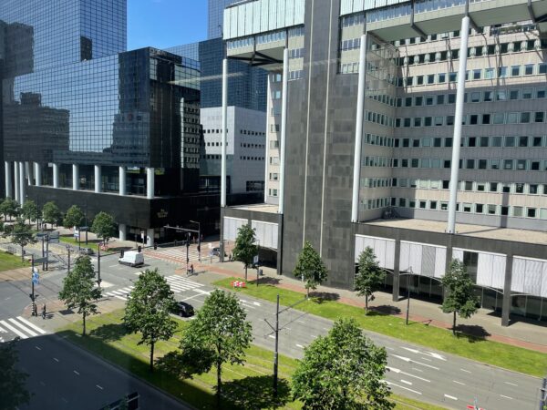 Straatbeeld van de Weena in Rotterdam met moderne kantoorgebouwen, verkeer en voetgangers op een zonnige dag.