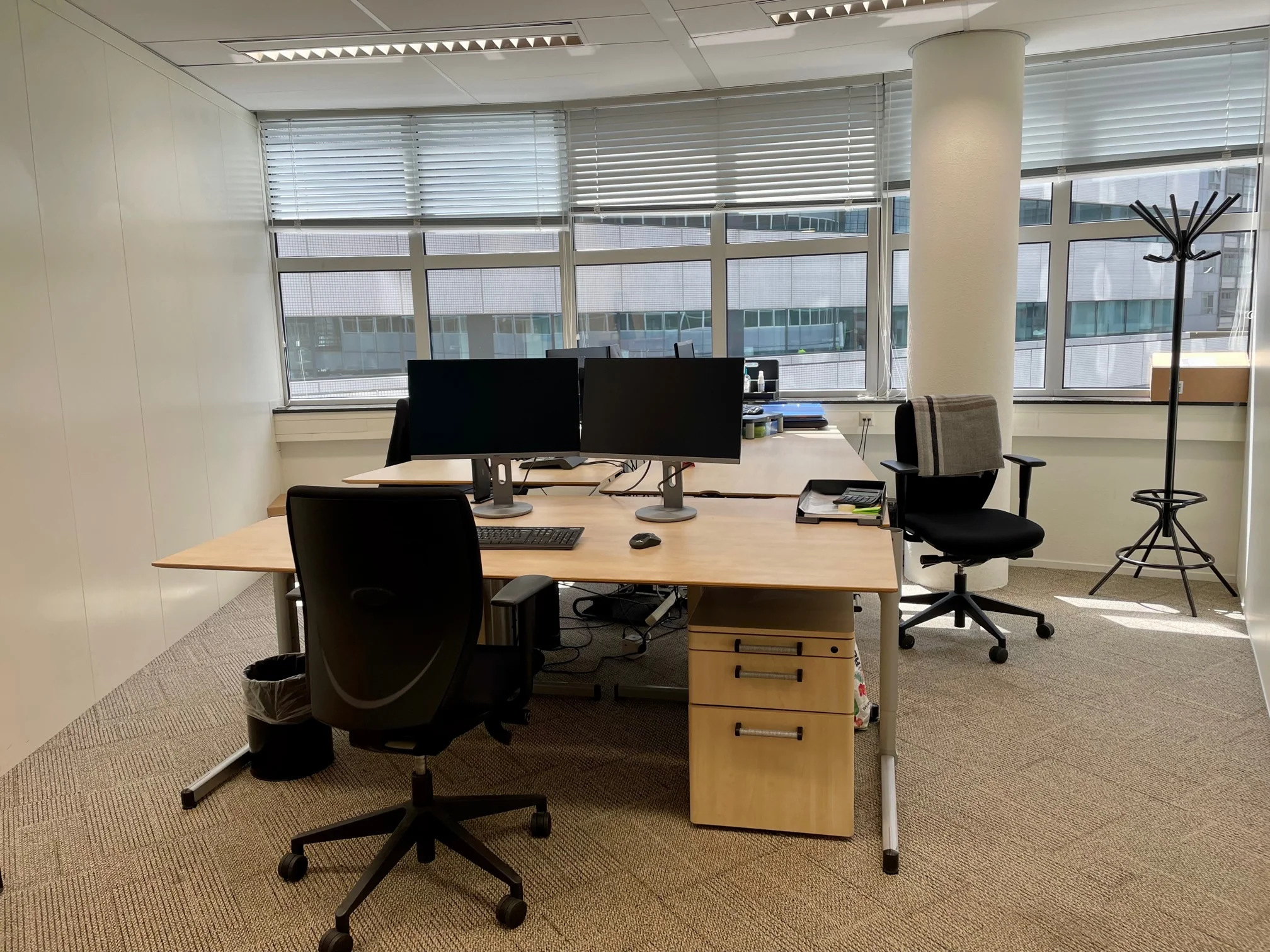 Kantoorruimte aan het Weena met twee bureaus, computerschermen, bureaustoelen en grote ramen met uitzicht op omliggende gebouwen.
