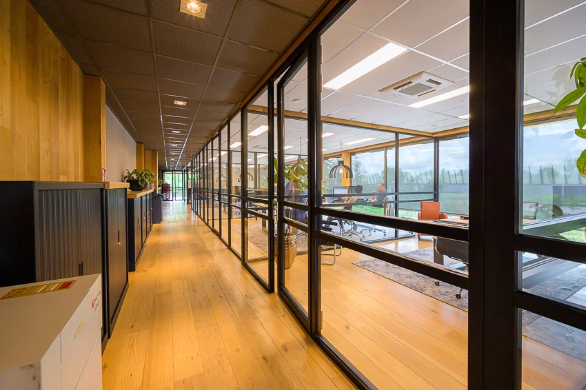 Gang met houten vloer en glazen wanden aan de Calandstraat, met zicht op een modern kantoor en vergaderruimte.