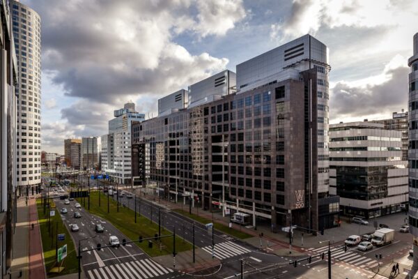 Drukke stadsstraat Weena in Rotterdam met moderne kantoorgebouwen en verkeer onder een deels bewolkte lucht.