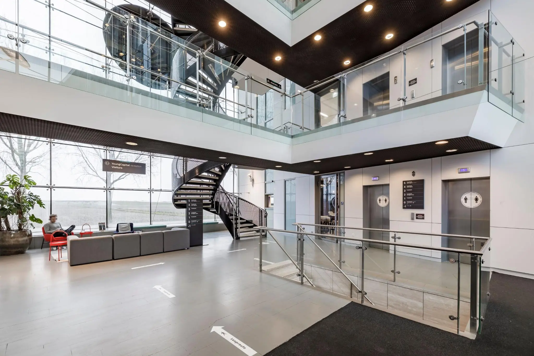 Moderne, lichte ontvangsthal van het kantoorpand aan de Siriusdreef met zitgedeelte, trap, liften en bewegwijzering naar YoungCapital.