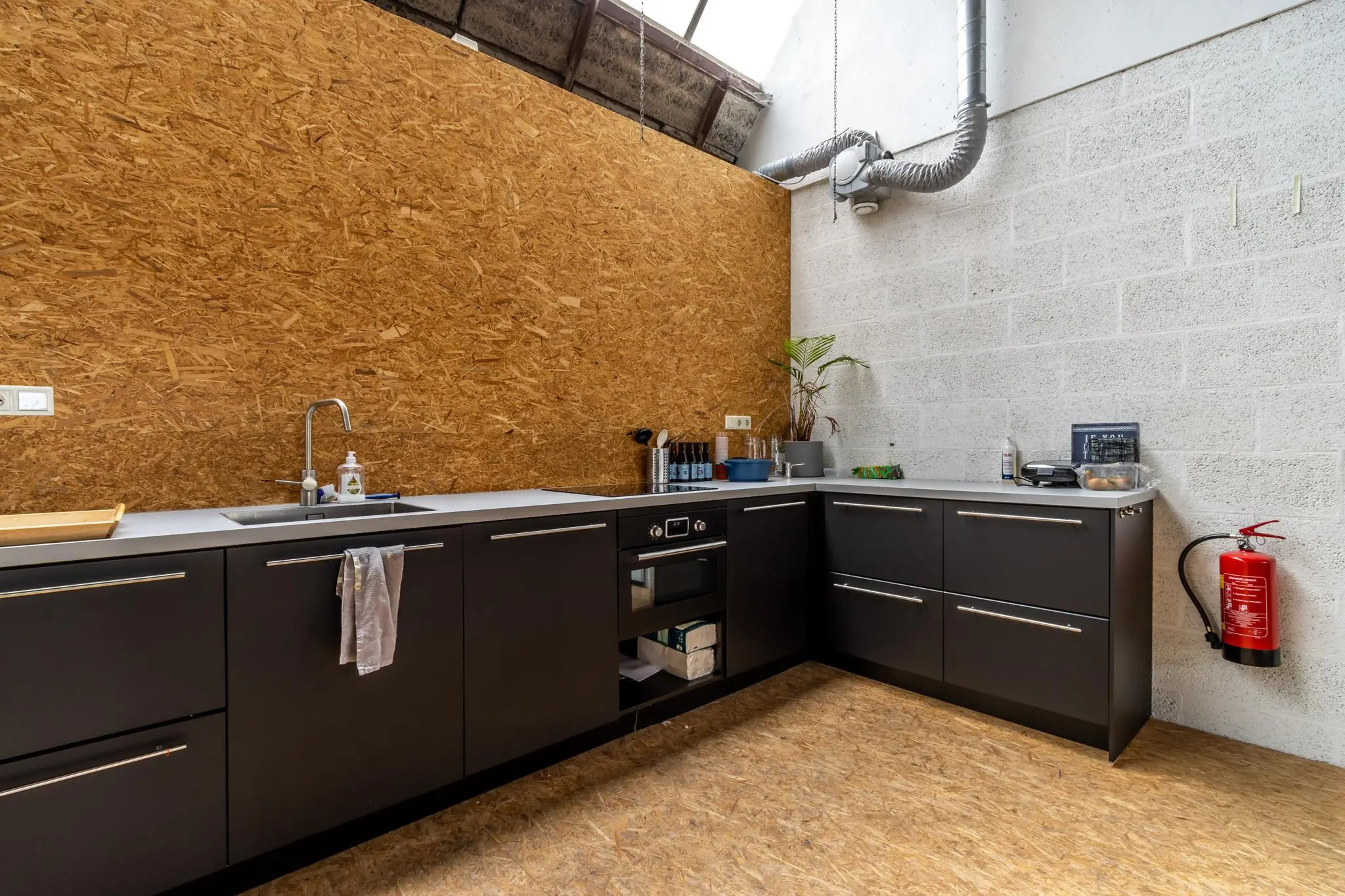 Moderne kitchenette met zwarte kastjes, een betonnen muur, OSB-houtafwerking en industriële ventilatie in een pand aan de Eendrachtlaan.