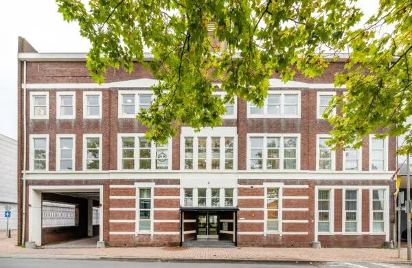 Voorgevel van een bakstenen kantoorpand aan de Stadionweg, gefotografeerd vanop straatniveau met boombladeren op de voorgrond.