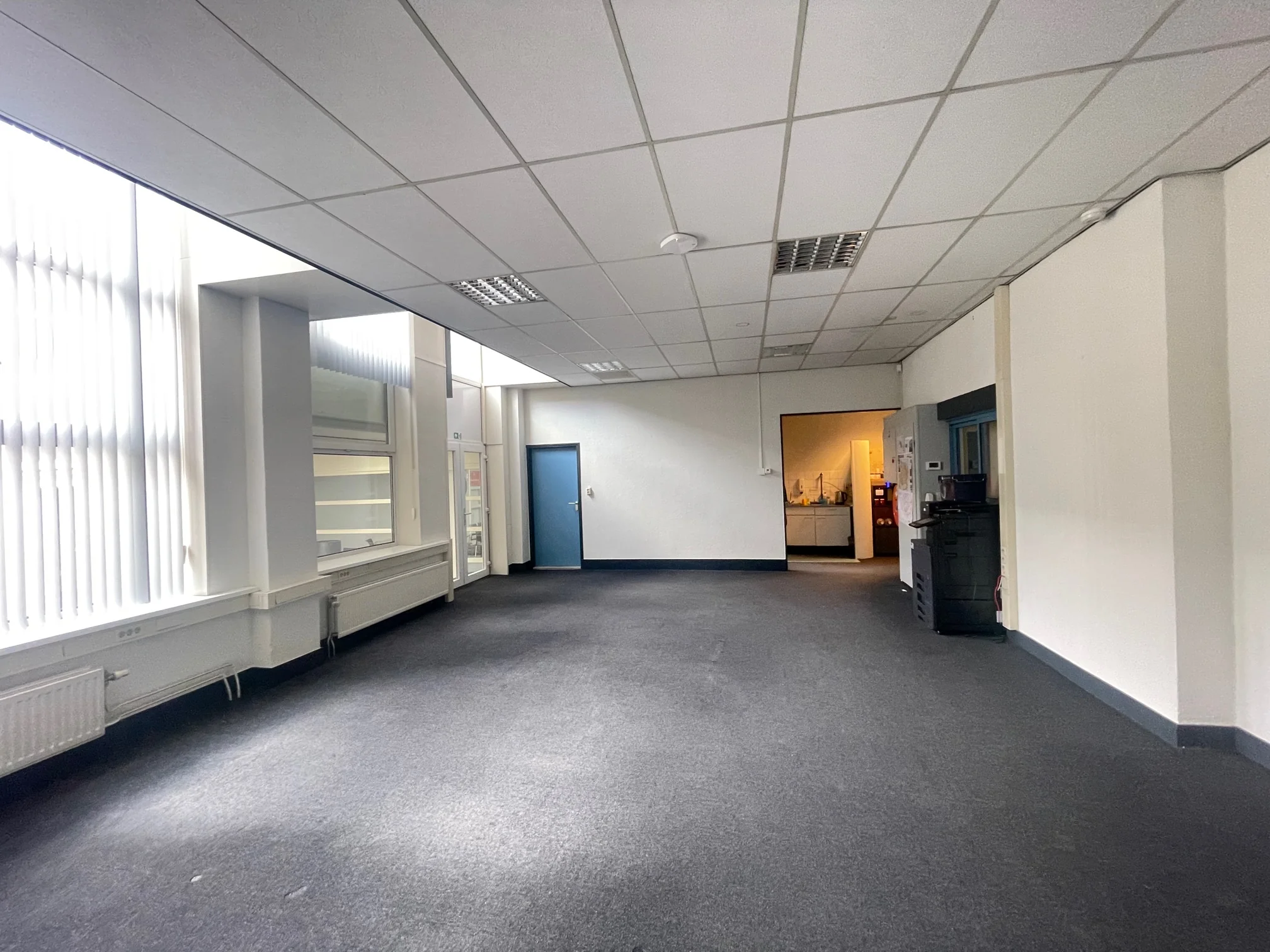 Lege kantoorruimte aan de Stadionweg met witte muren, systeemplafond en grote ramen met verticale lamellen.