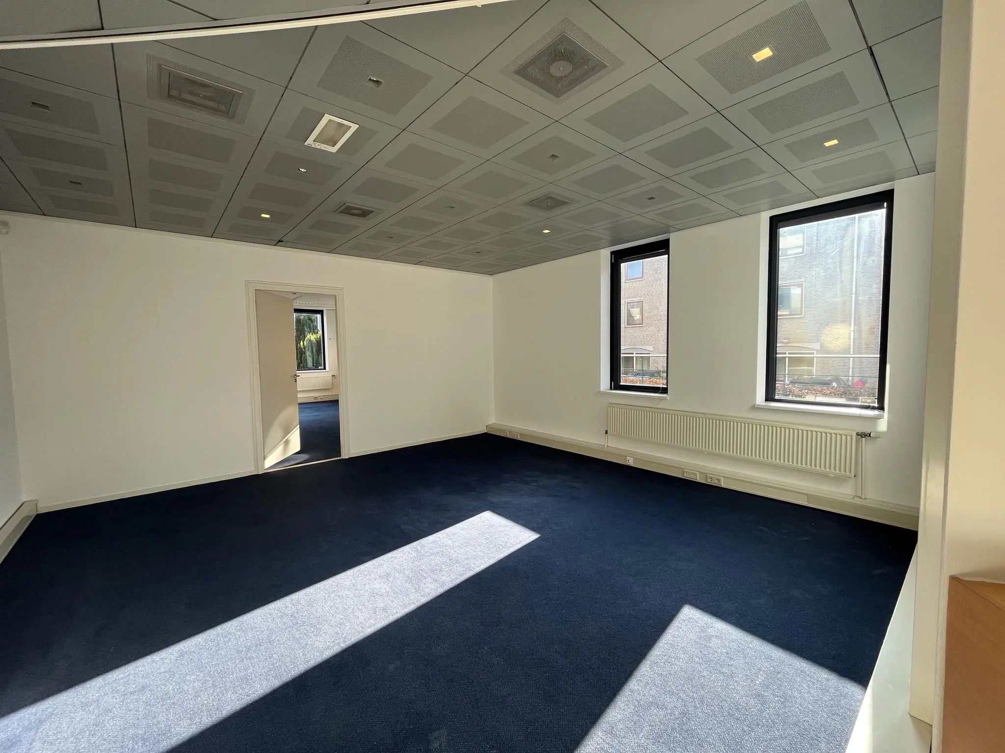 Lege kantoorruimte aan de Amersfoortseweg met donkerblauwe vloerbedekking, witgeverfde muren en twee grote ramen die uitkijken op een naastgelegen gebouw.