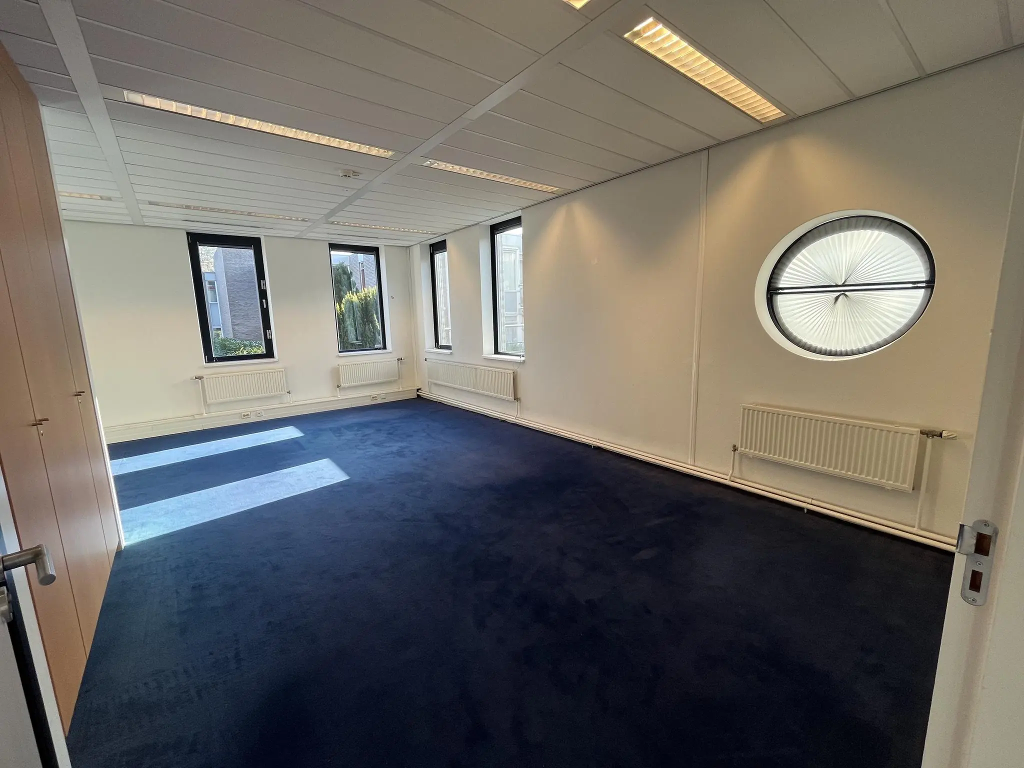 Licht kantoorruimte aan de Amersfoortseweg met donkerblauwe vloerbedekking, meerdere ramen en een ronde designradiator.