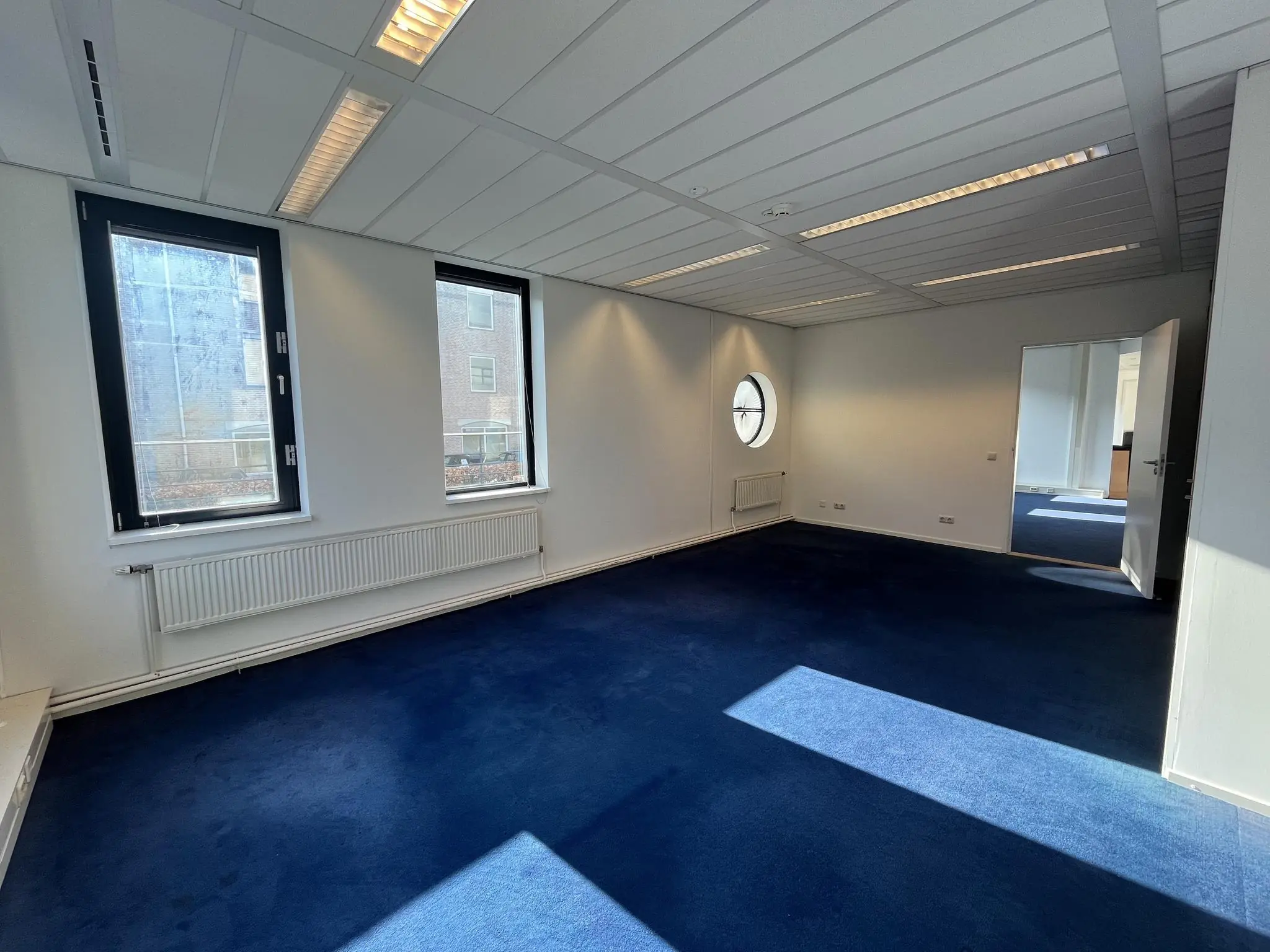 Lichte, lege kantoorruimte aan de Amersfoortseweg met blauwe vloerbedekking en grote ramen.