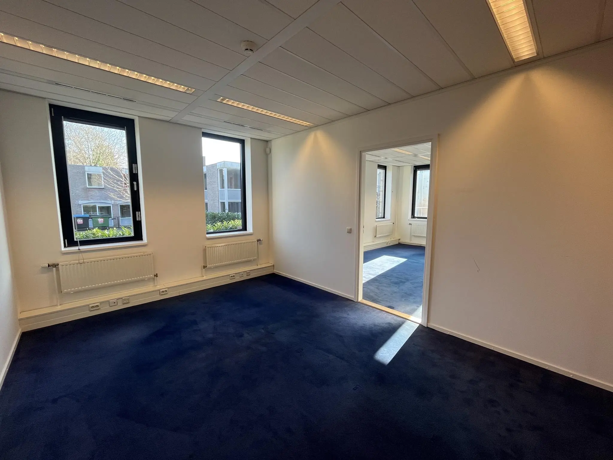 Een leeg kantoorpand aan de Amersfoortseweg met donkerblauw tapijt en grote ramen die uitzicht bieden op de omgeving.