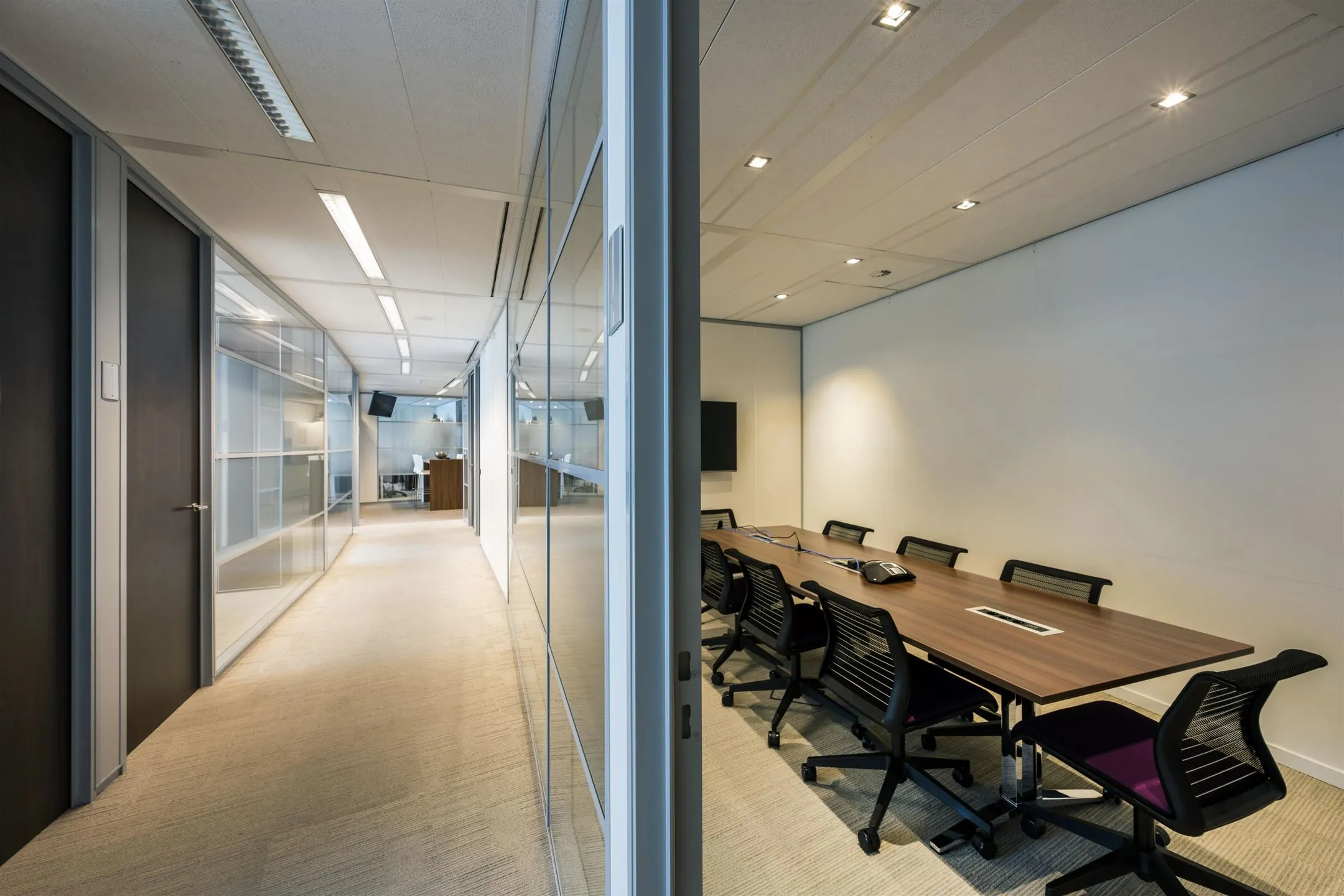 Moderne kantoorruimte met een gang en een vergaderruimte met een lange tafel en meerdere stoelen.