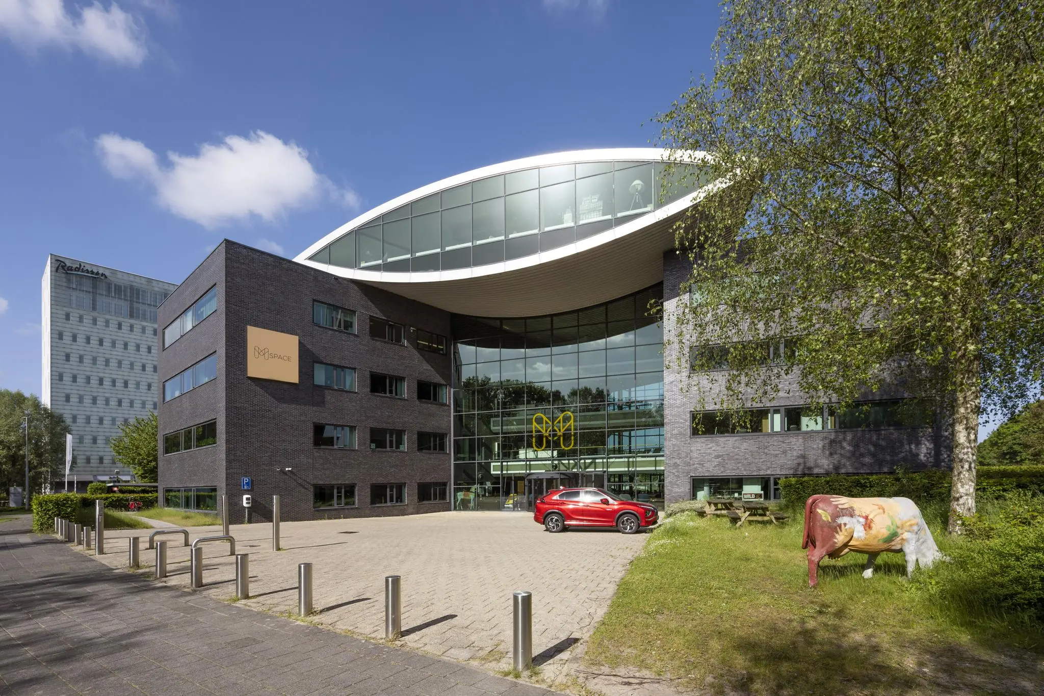 Modern kantoorgebouw aan de Prof. W.H. Keesomlaan met glazen gevel, kunstkoe op het gras en een rode auto op de oprijlaan.