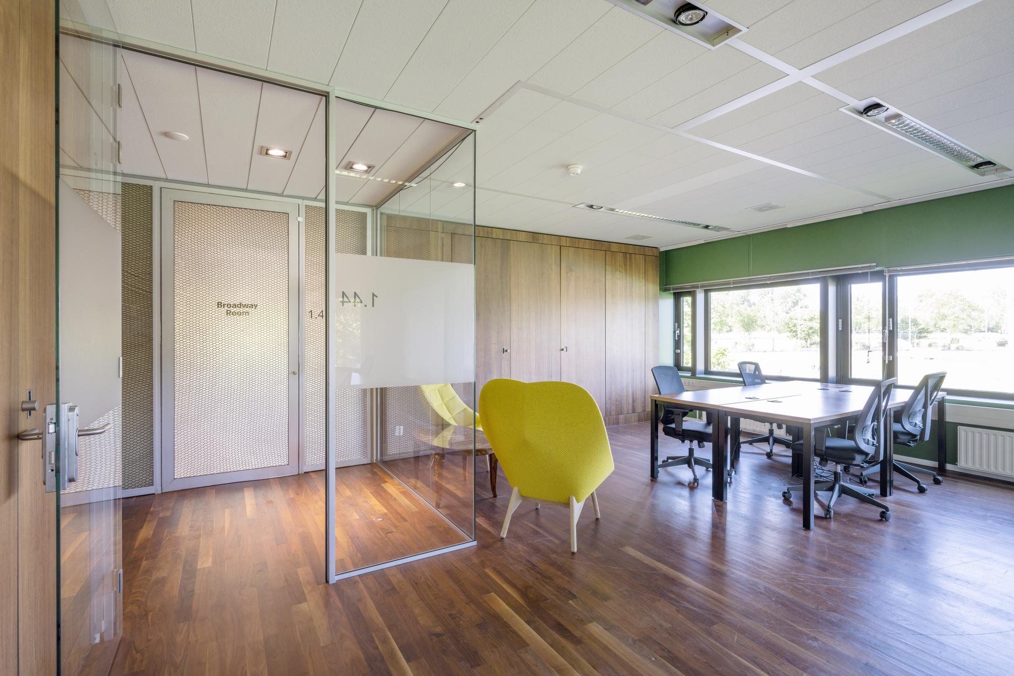 Moderne vergaderruimte aan de Prof. W.H. Keesomlaan met glazen wanden, houten vloer, werkplekken en een opvallende gele stoel.