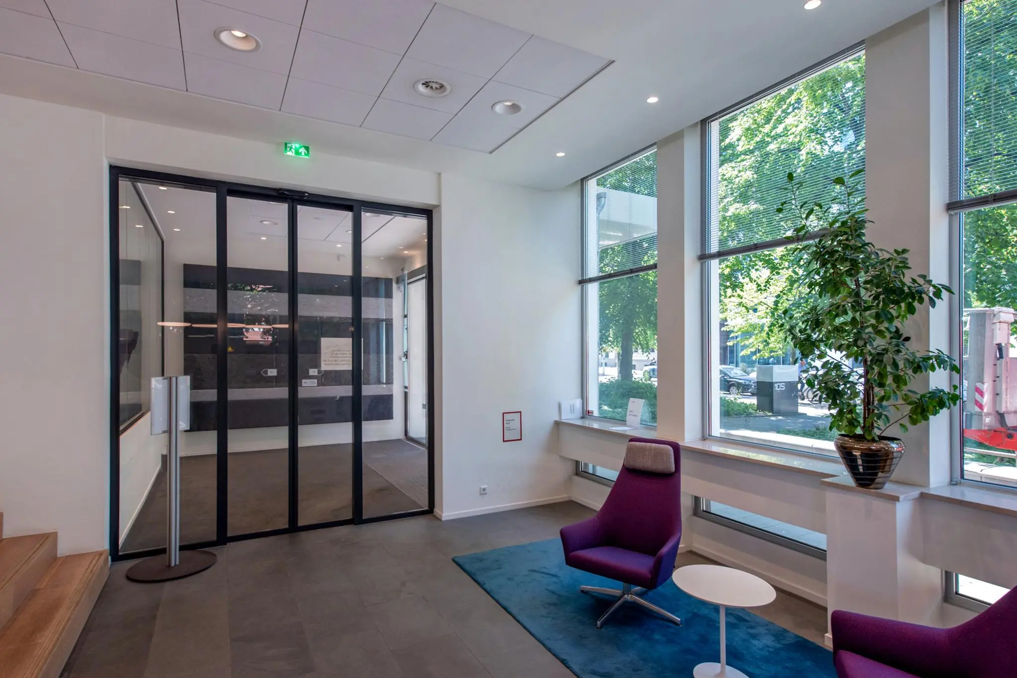Moderne ontvangsthal aan de Bezuidenhoutseweg met glazen deuren, paarse stoelen en grote ramen met uitzicht op de straat.