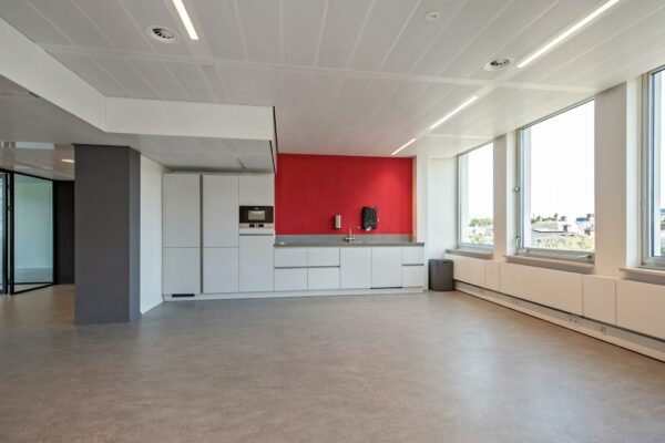 Moderne kantoorpantry aan de Bezuidenhoutseweg met witte kasten, een rode accentmuur en grote ramen die veel daglicht binnenlaten.
