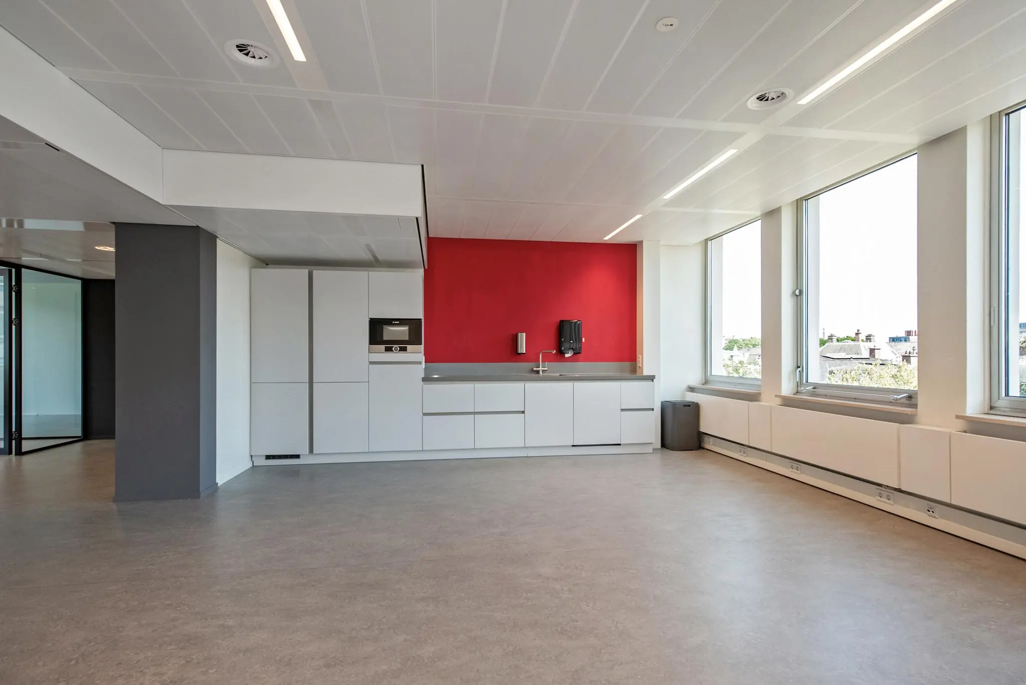 Moderne kantoorpantry aan de Bezuidenhoutseweg met witte kasten, een rode accentmuur en grote ramen die veel daglicht binnenlaten.