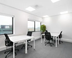 Moderne, lichte kantoorruimte met vier bureaus, ergonomische stoelen en grote ramen met uitzicht op kantoorgebouwen.