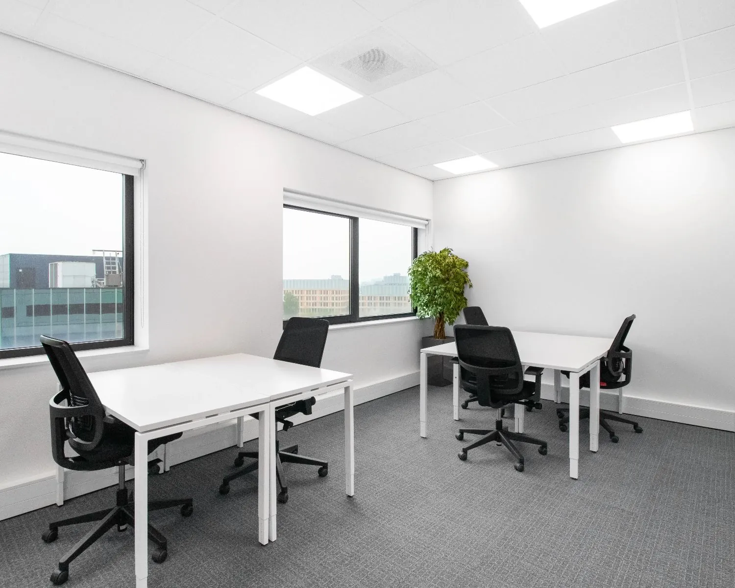 Moderne, lichte kantoorruimte met vier bureaus, ergonomische stoelen en grote ramen met uitzicht op kantoorgebouwen.