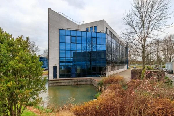 Modern kantoorgebouw aan de Vlierbaan met spiegelende ramen, gelegen aan een vijver en omringd door bomen en struiken.