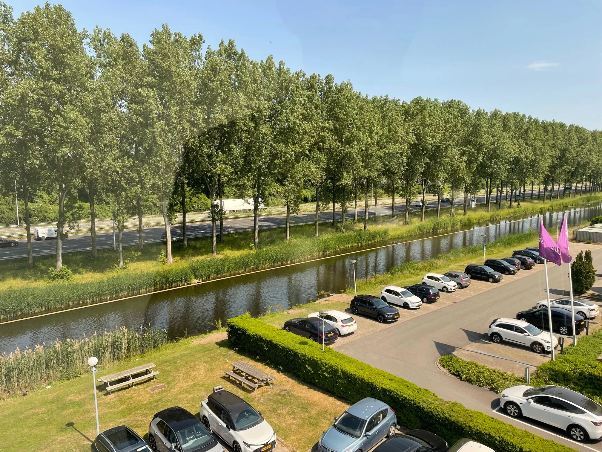 Parkeerterrein aan de Vlierbaan met geparkeerde auto's, een watergang en een rij bomen langs een snelweg op de achtergrond.