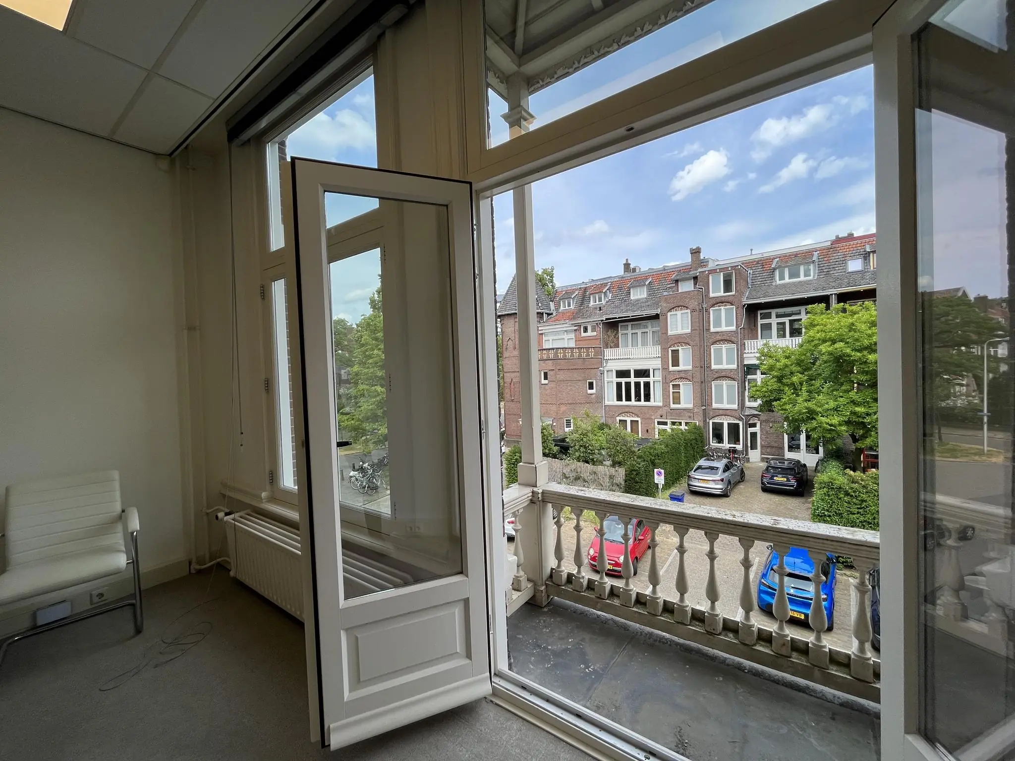 Openstaand raam in een kamer aan de Ramstraat met uitzicht op klassieke woonhuizen en geparkeerde auto's.