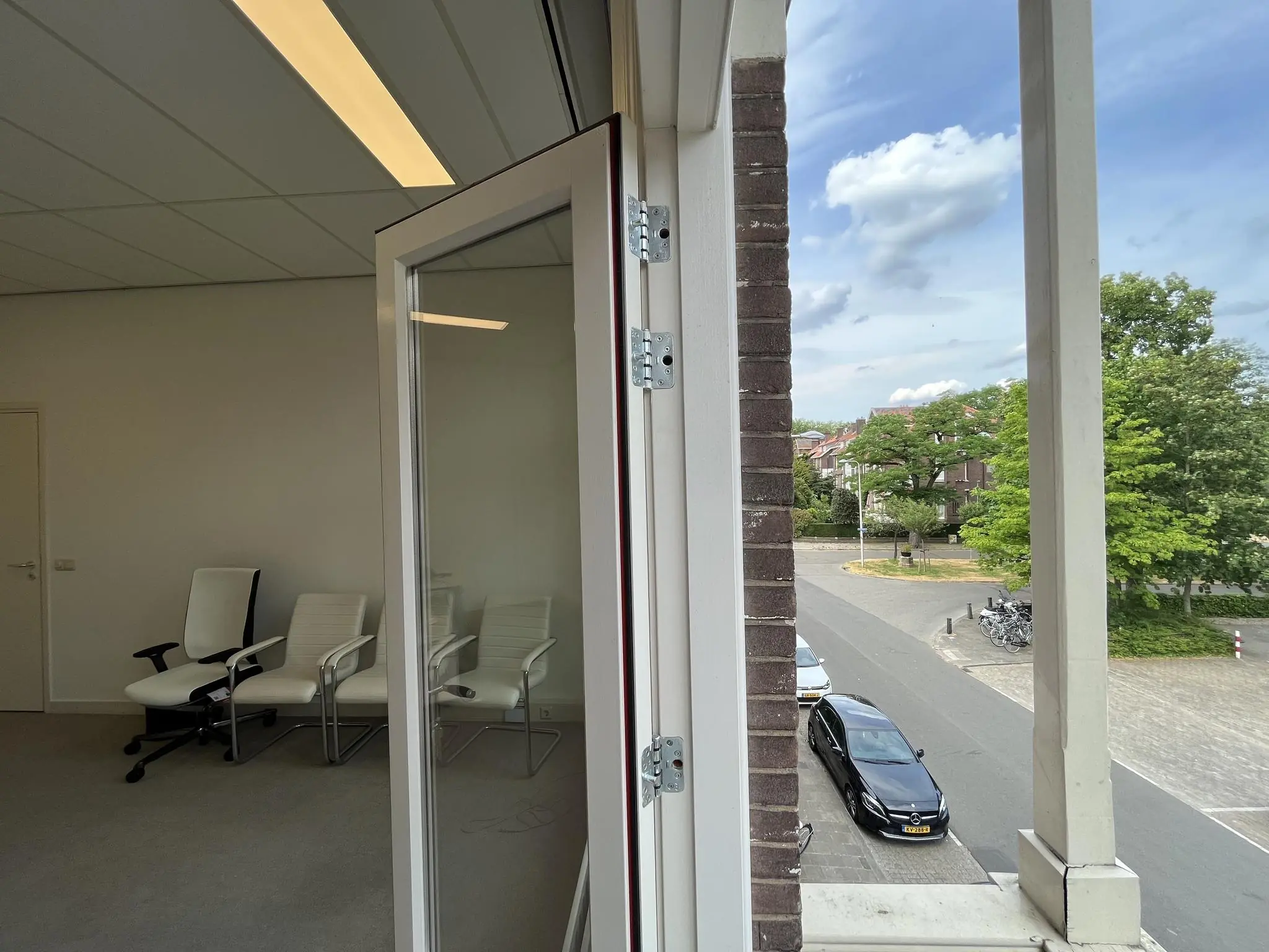 Uitzicht vanuit een open raam op de Ramstraat, met zicht op een rustige straat en enkele geparkeerde auto's, en binnenin een kantoorruimte met drie witte stoelen.