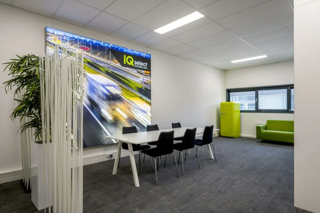 Moderne vergaderruimte met witte tafel, zwarte stoelen, groene bank en koelkast, en een grote wandfoto van snelwegverkeer met het logo van IQ Select.