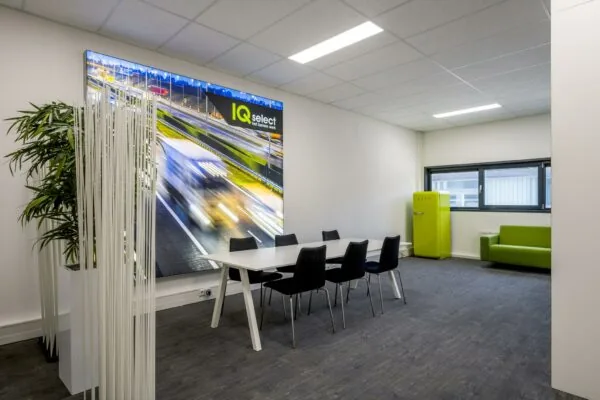 Moderne vergaderruimte met witte tafel, zwarte stoelen, groene bank en koelkast, en een grote wandfoto van snelwegverkeer met het logo van IQ Select.